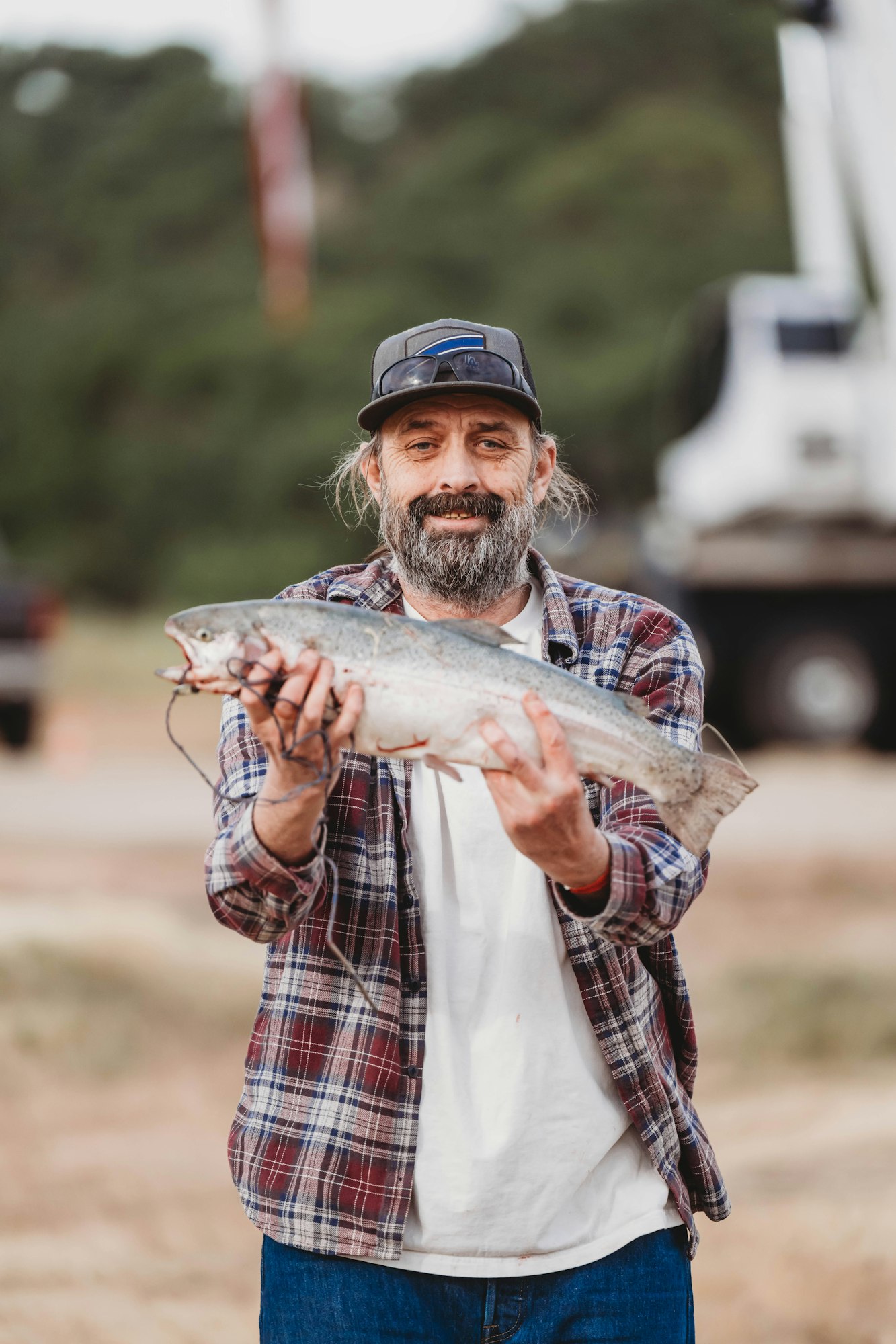A person holding a fish, wearing a plaid shirt, white t-shirt, and a hat, standing outdoors.