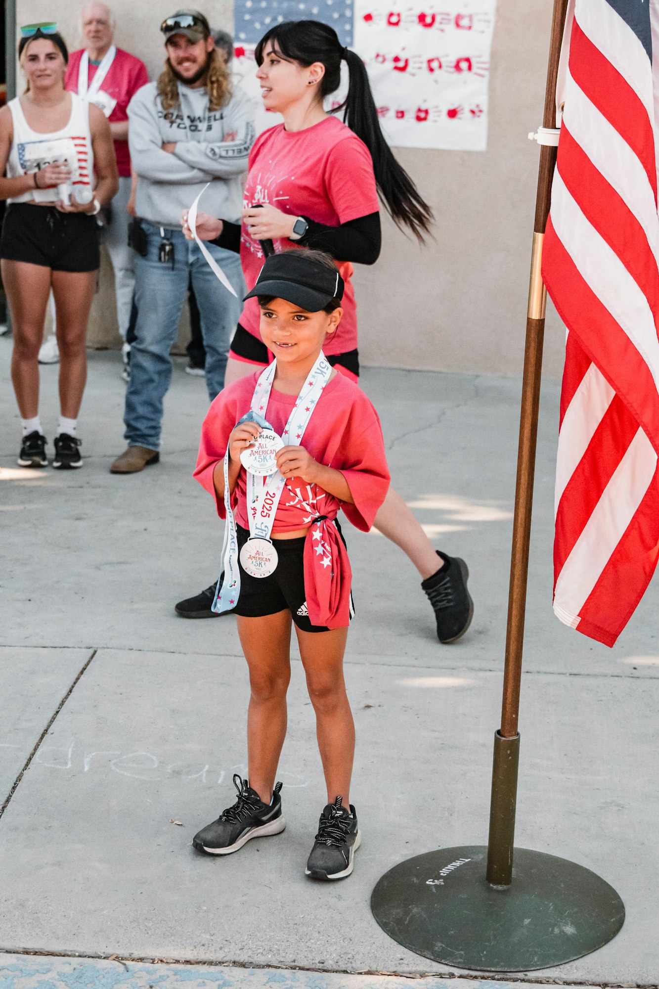 A young girl holds medals, standing by an American flag, with people in the background.
