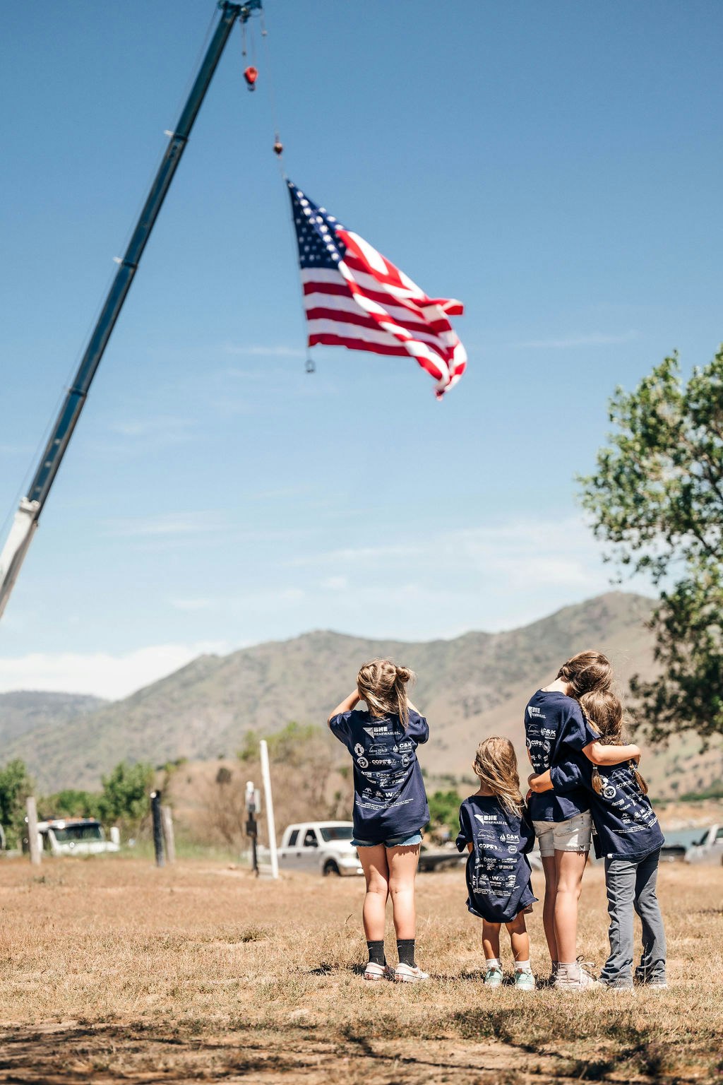 Four children watching an American flag hoisted by a crane against a clear sky.