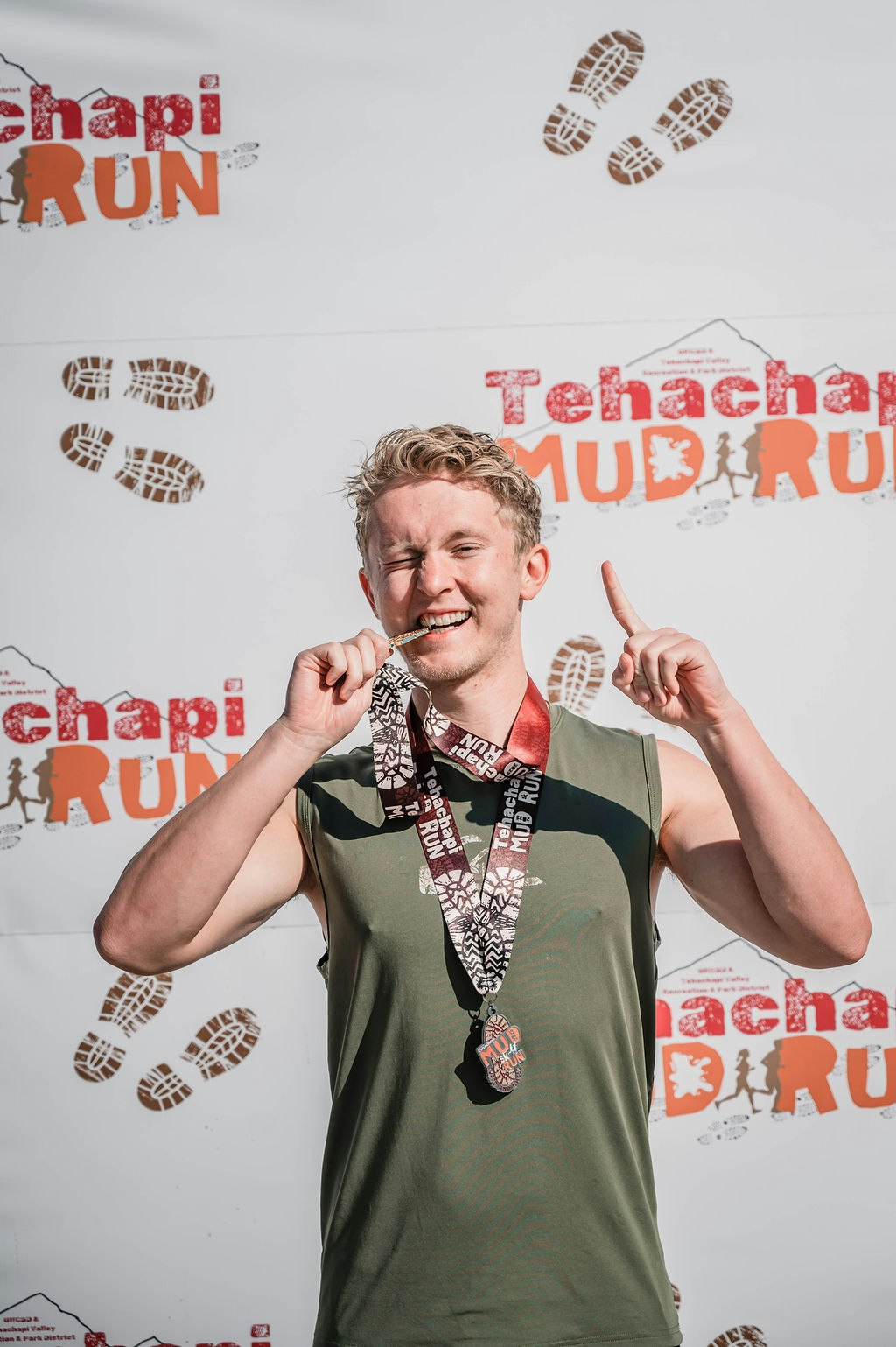 Person celebrating with a medal in front of a "Tehachapi Mud Run" banner.