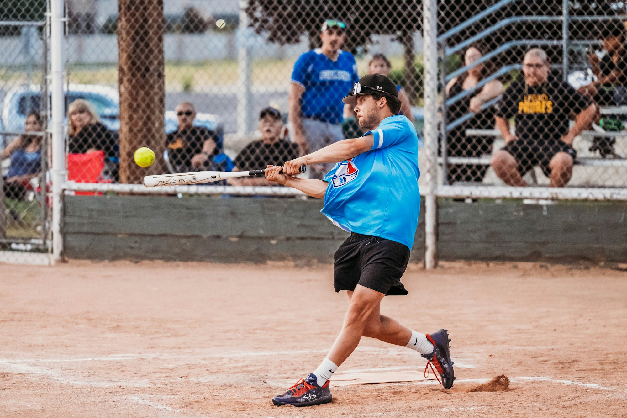 A softball player swings the bat at a pitch with spectators in the background.