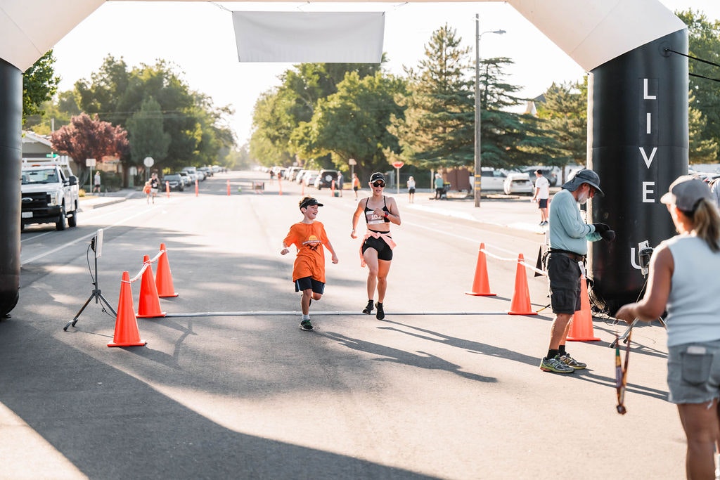 People finishing a race, crossing a finish line with orange cones, a bright street, and trees in the background.
