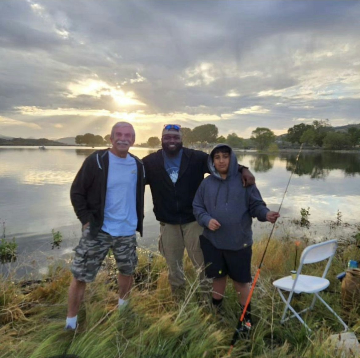 Three people by a lakeside at sunset, with fishing gear and a folding chair.