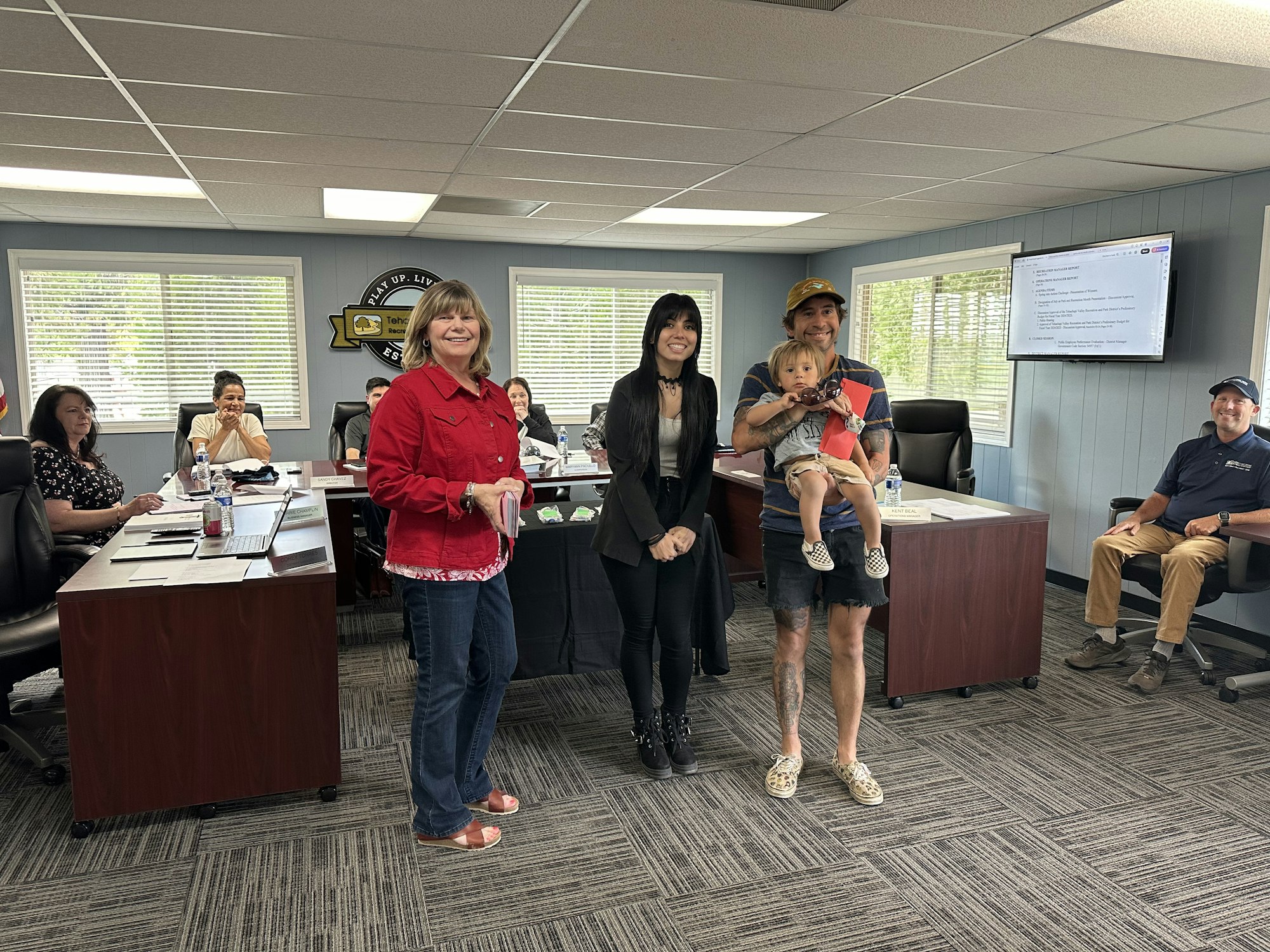 People in a meeting room with one holding a toddler, smiling, possibly a casual or community gathering.