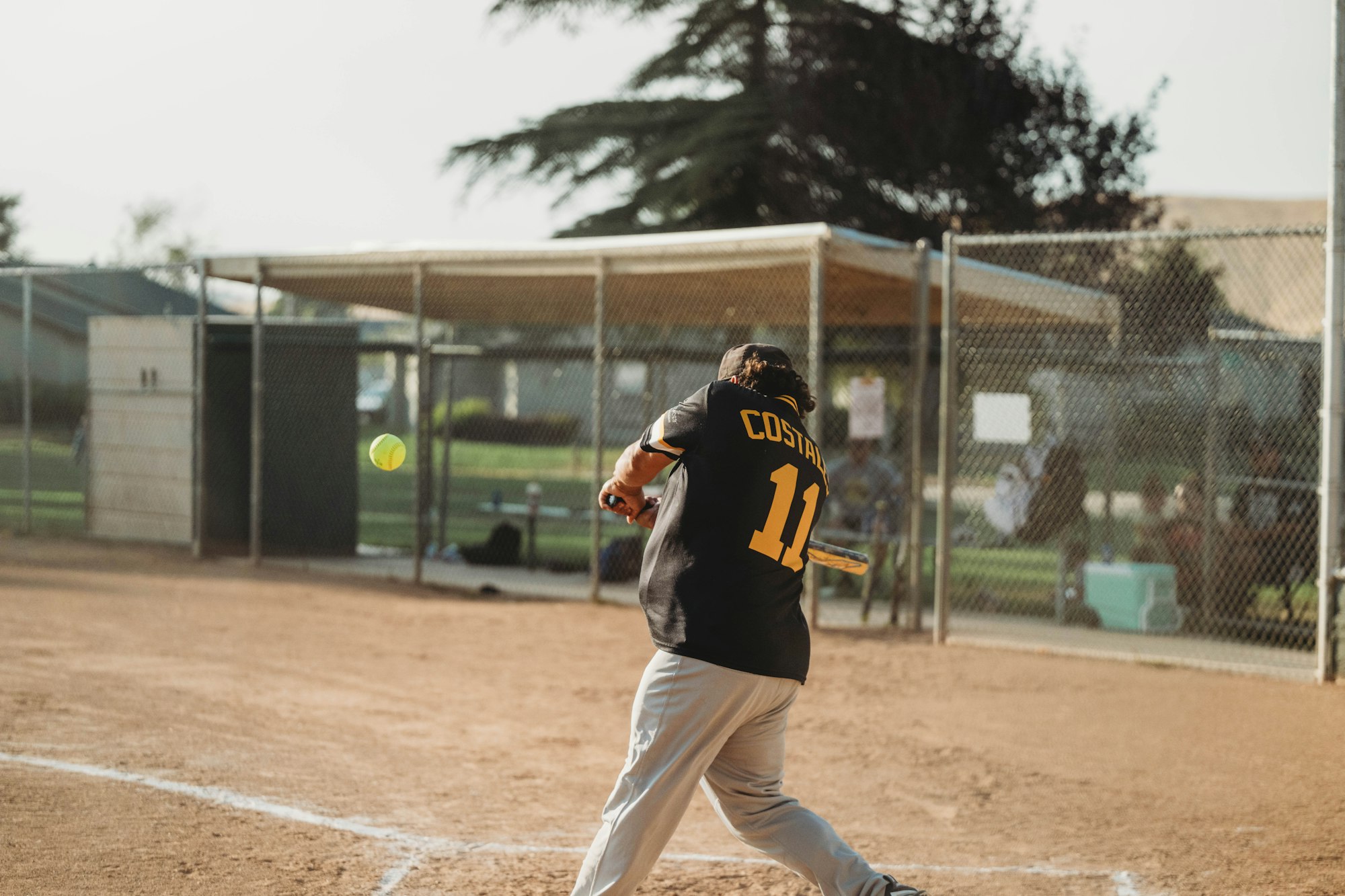 A player is swinging a bat at a softball during a game or practice on a sandy field, wearing a jersey with the name "COSTAL."