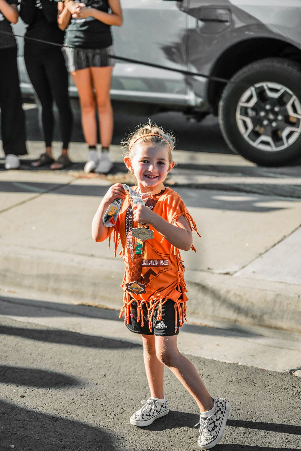 A smiling child holds medals, wearing an orange fringed shirt and sneakers, standing on a street with people in the background.