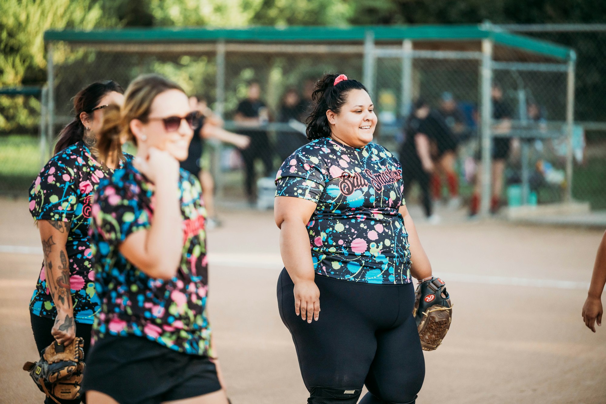 Women in colorful sports attire on a softball field, one smiling and holding a glove.