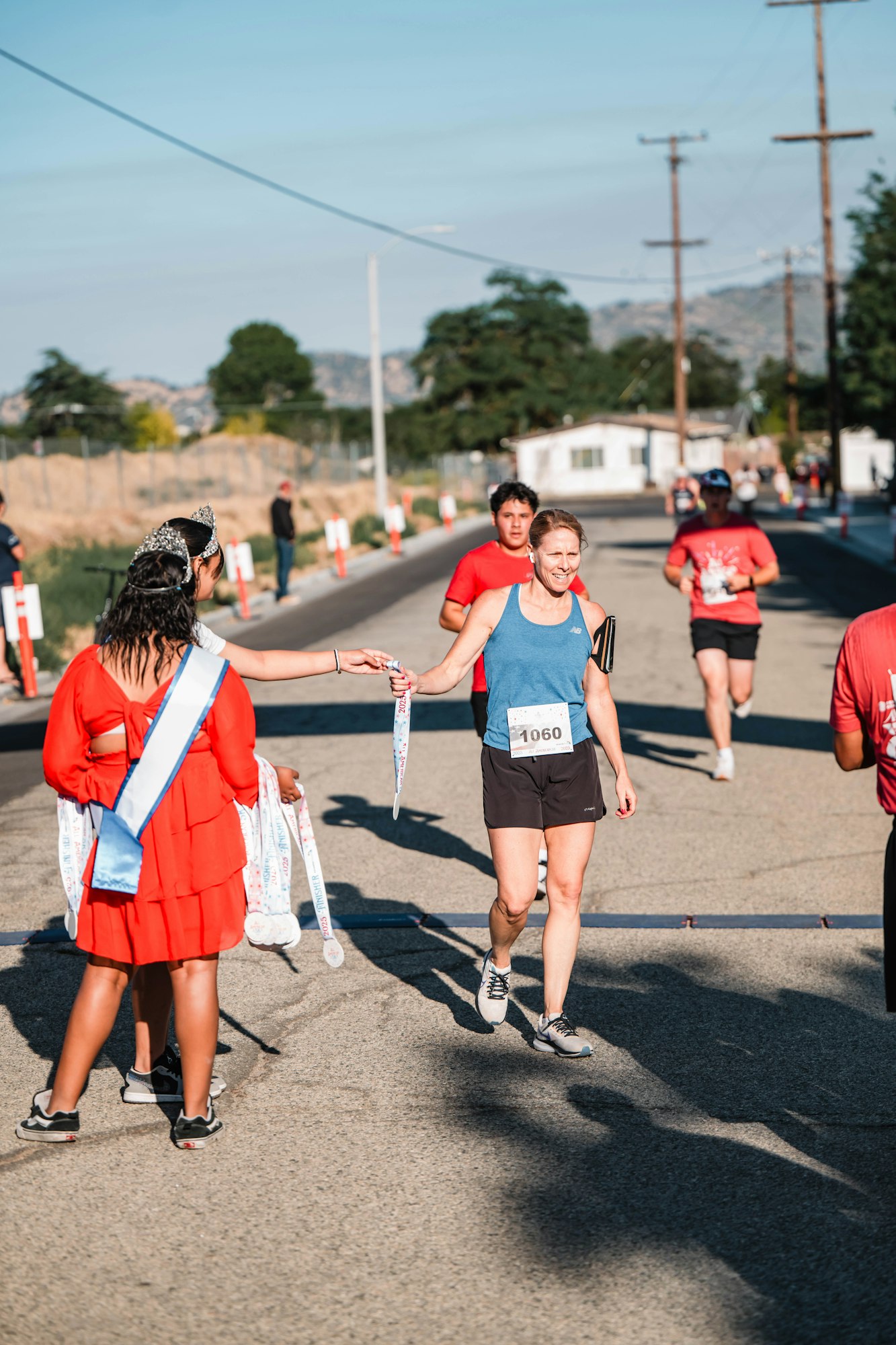 A runner receives a medal from a person in a red dress and sash after finishing a race. Other participants are in the background.