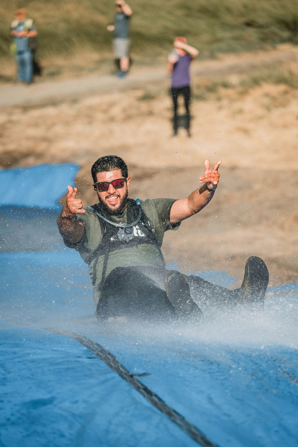Person sliding down a wet, blue tarp outdoors, smiling and pointing, with two people in the background.