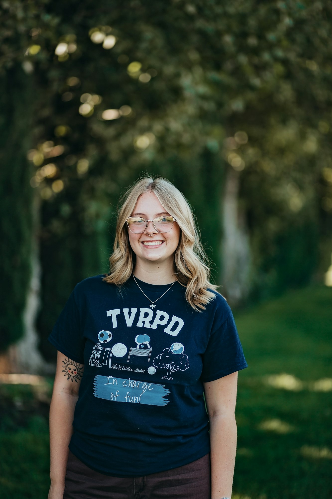 A smiling person in a T-shirt that says "TVRPD ... in charge of fun!" standing in front of trees.