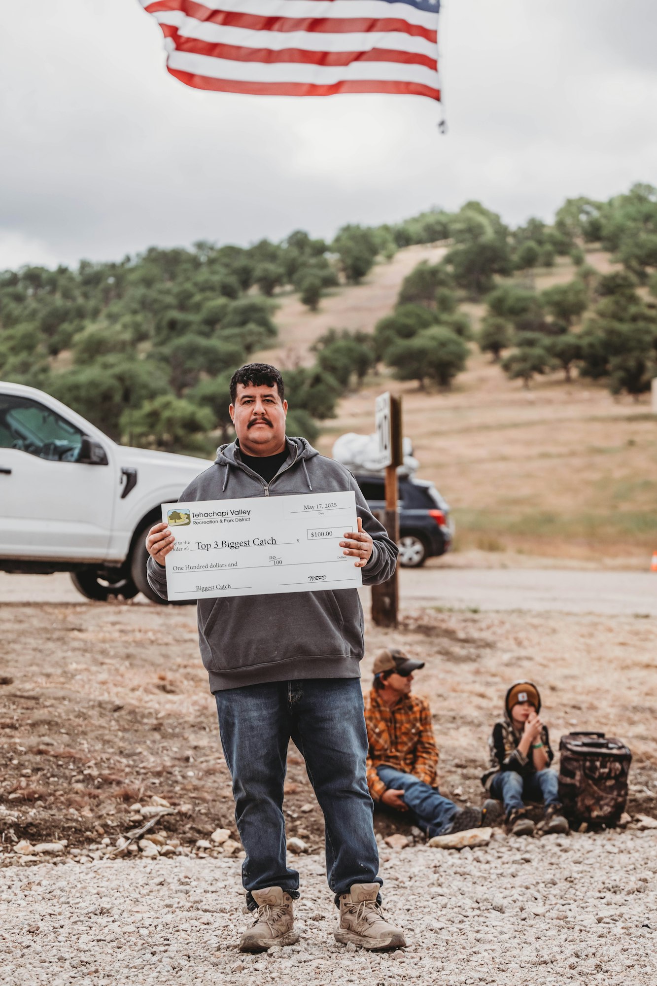 Man holding a large check for "Top 3 Biggest Catch," standing outdoors under an American flag, with others sitting in the background.