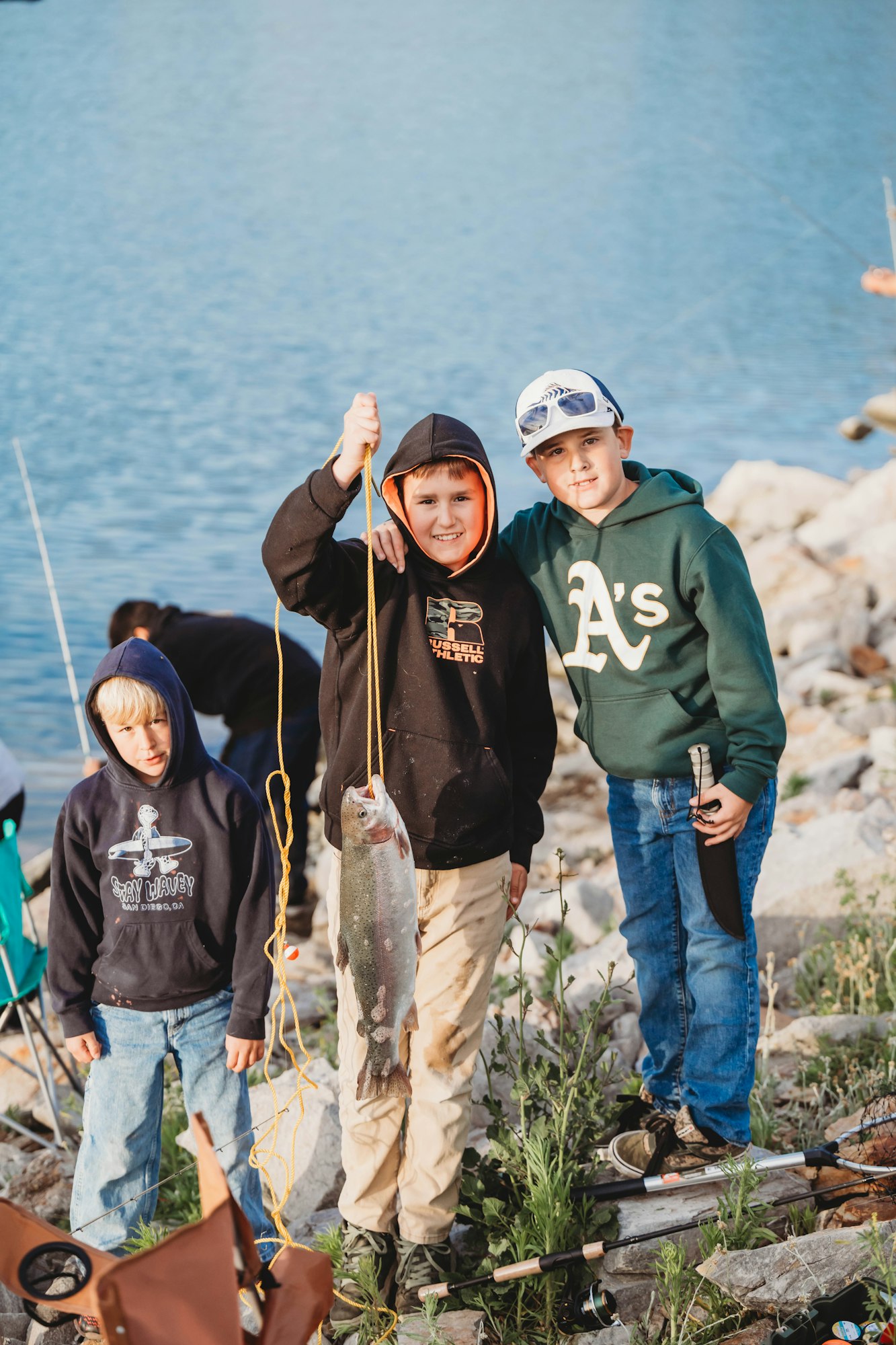 Three boys by a lake, one holding a large fish with a rope, smiling. Rocks and fishing gear are visible.