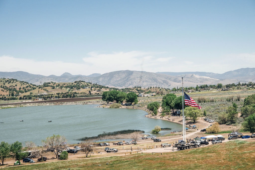 A serene lakeside landscape with an American flag, boats on water, parked cars, and distant hills under a clear sky.