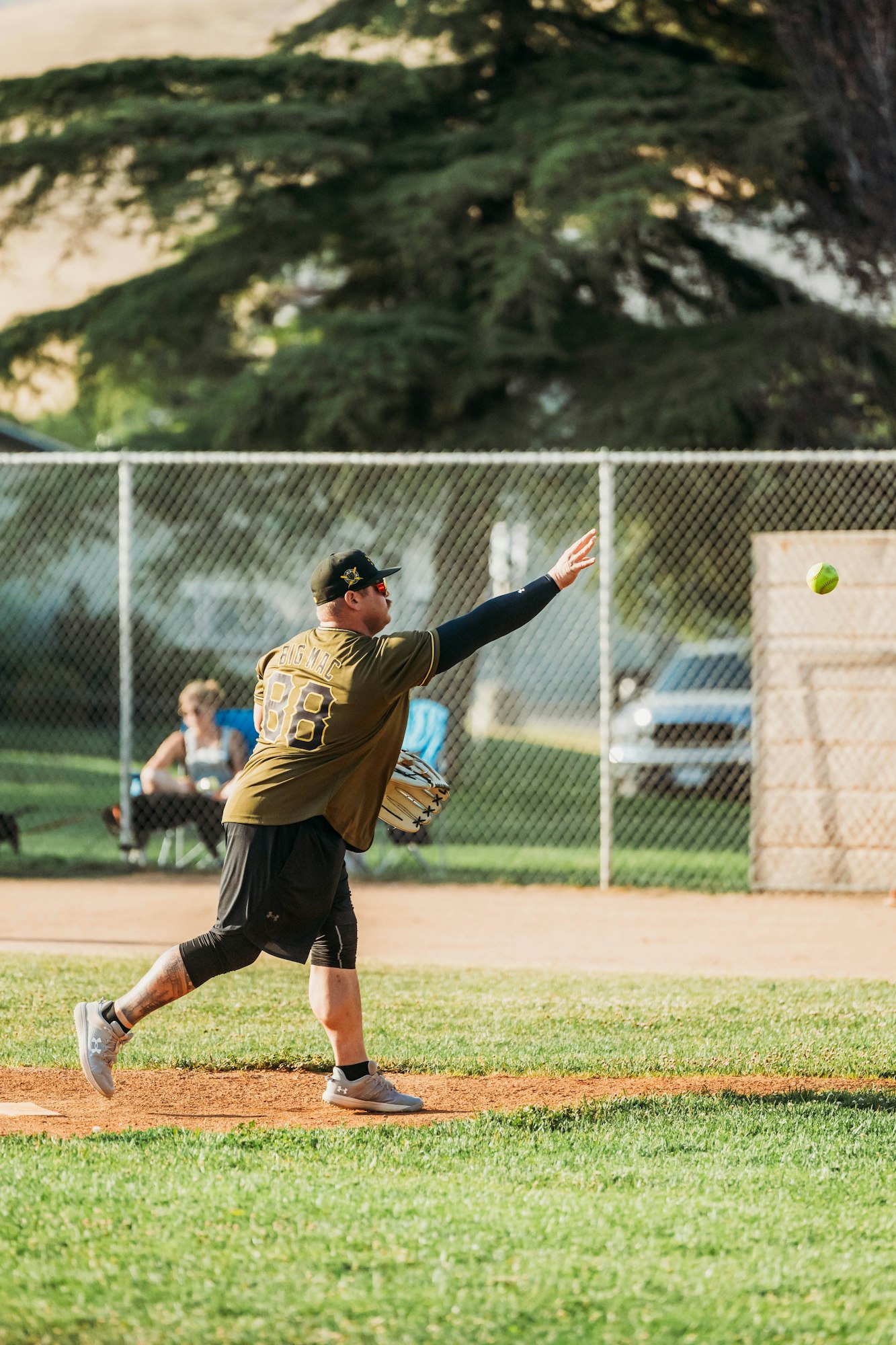 A baseball player pitching on a sunny day, trees and a fence in the background.