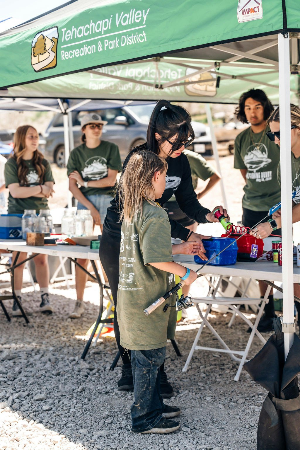 A group of people under a branded tent, likely at an outdoor event, with a child in front receiving guidance or an item.