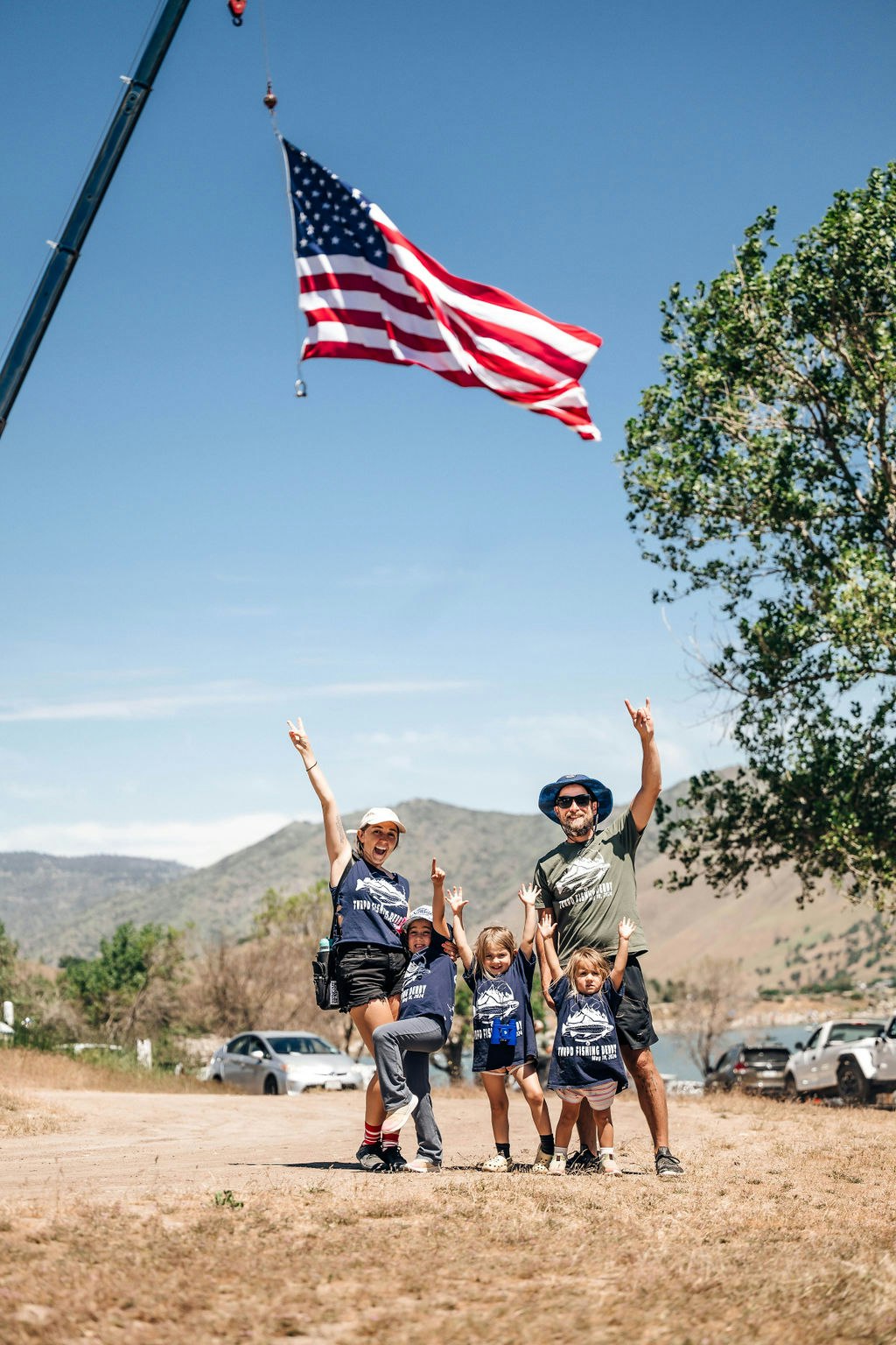 A family posing joyfully under a large American flag suspended by a crane, outdoors on a sunny day.