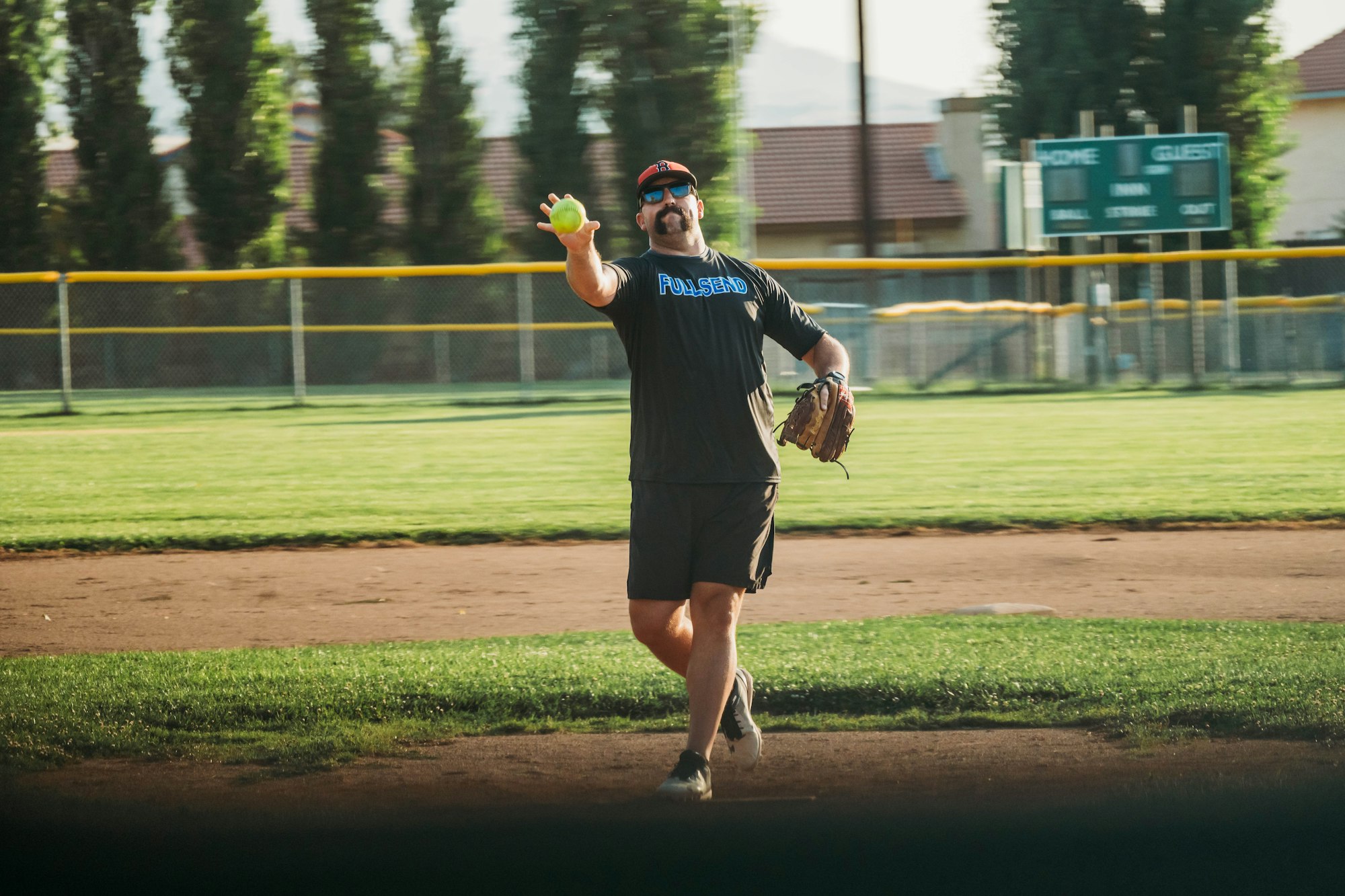 Person playing softball on a field, wearing sunglasses and a cap, about to throw a ball. Baseball diamond in the background.