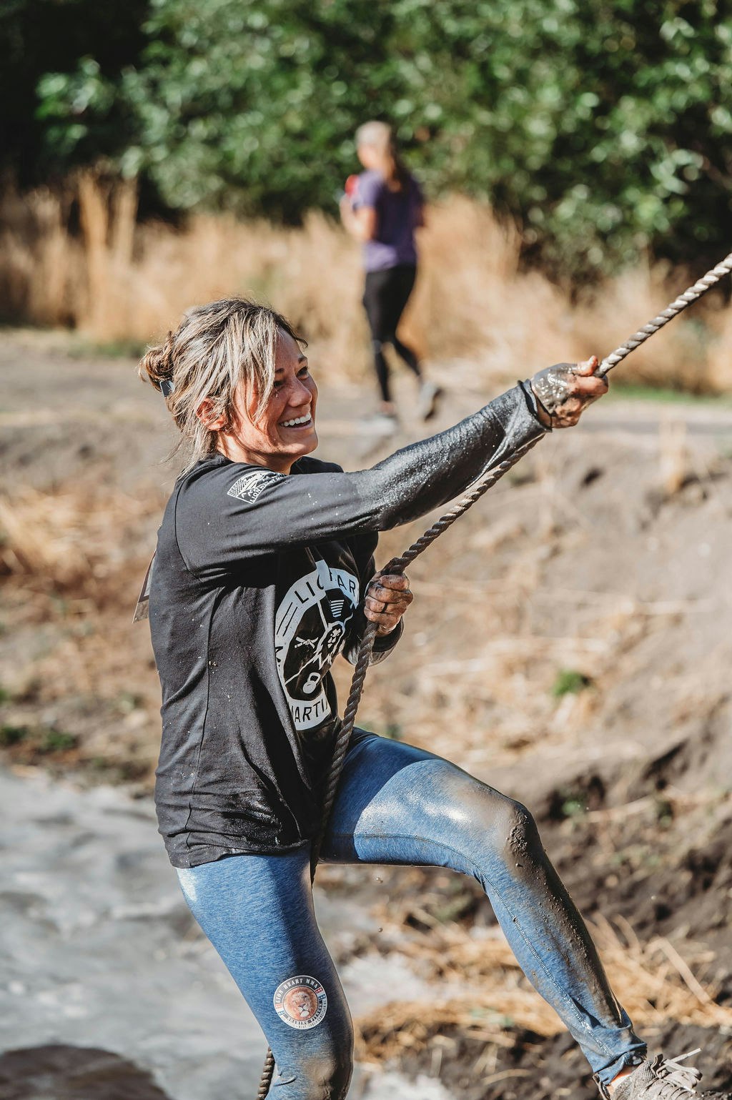 A woman smiling while climbing a muddy rope during an outdoor obstacle course.