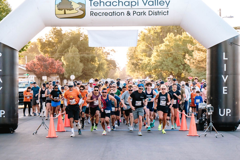 A group of runners starting a race under a Tehachapi Valley banner with trees in the background.