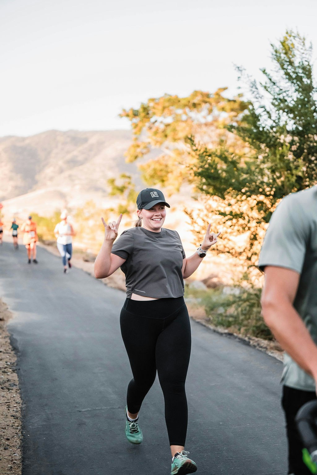 A person running on a paved path with a joyful expression, wearing a cap, and making hand gestures, with others running in the background.