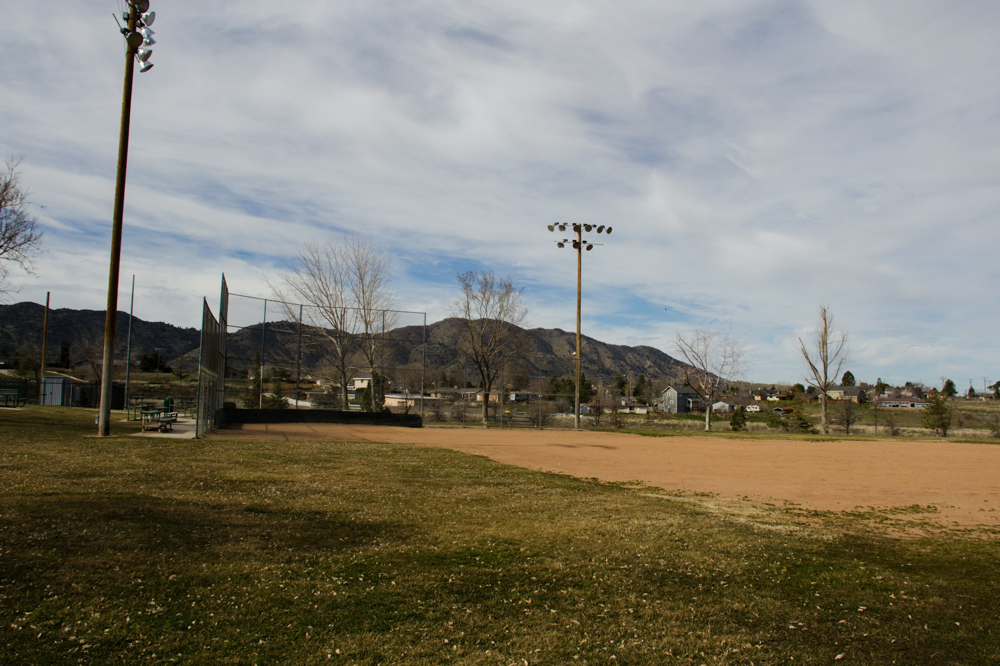 A baseball field with grass, dirt infield, fence, lighting, surrounding trees, and distant hills under a partly cloudy sky.