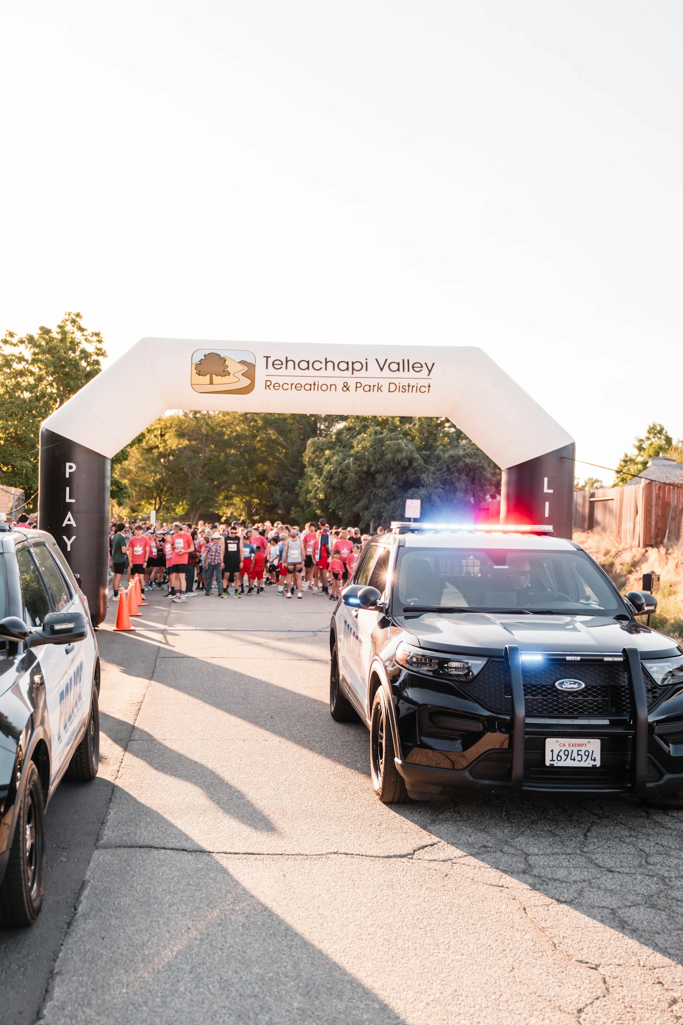Runners gather at a start line under an archway for Tehachapi Valley Recreation, near police cars with lights on.