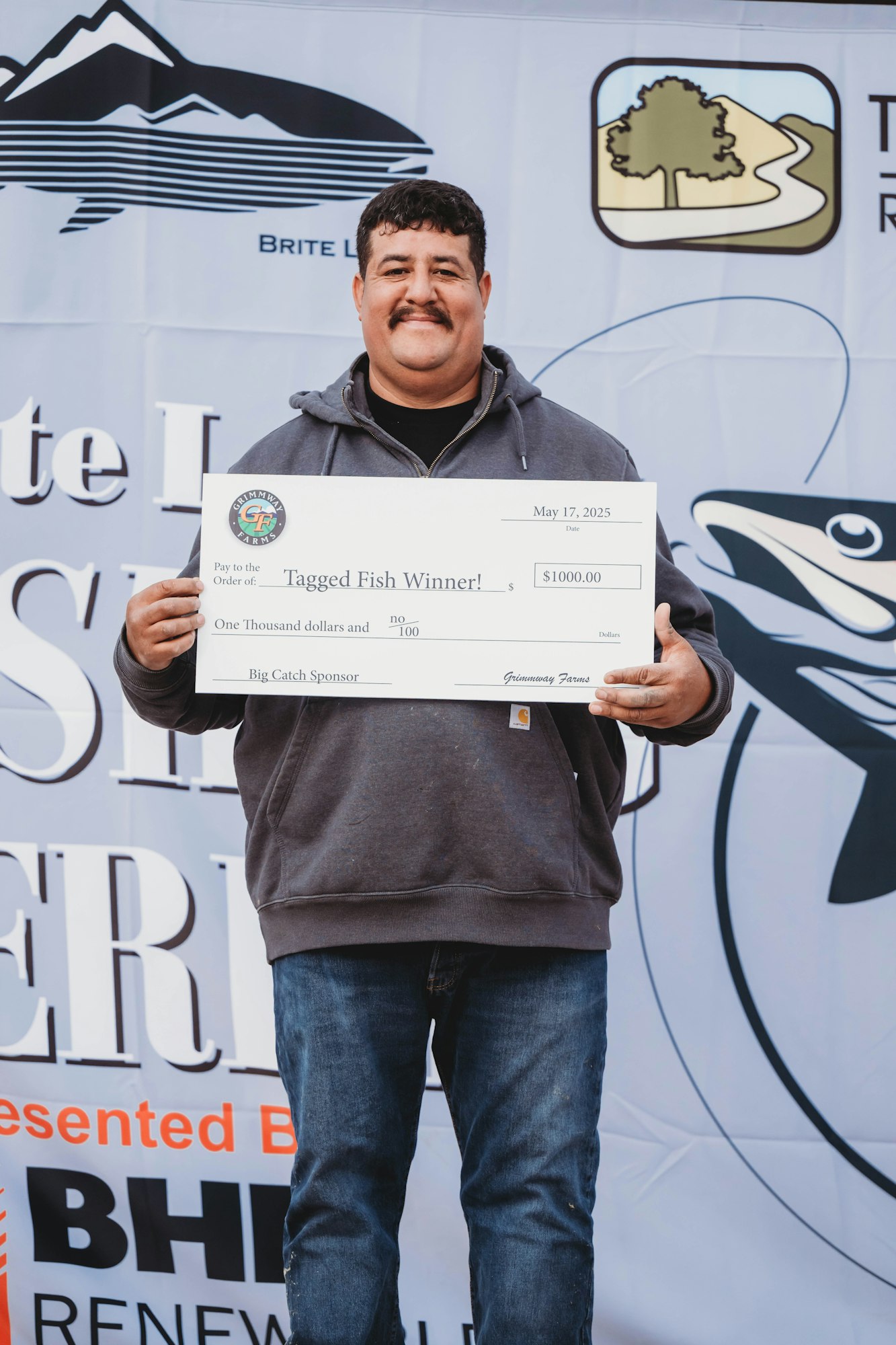 A person holding a large check for $1000, awarded as "Tagged Fish Winner!”