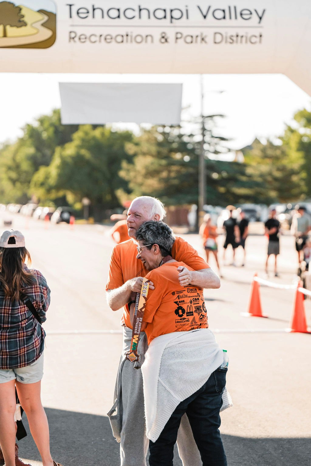 People in orange shirts hugging at a race finish line under a "Tehachapi Valley Recreation & Park District" banner.