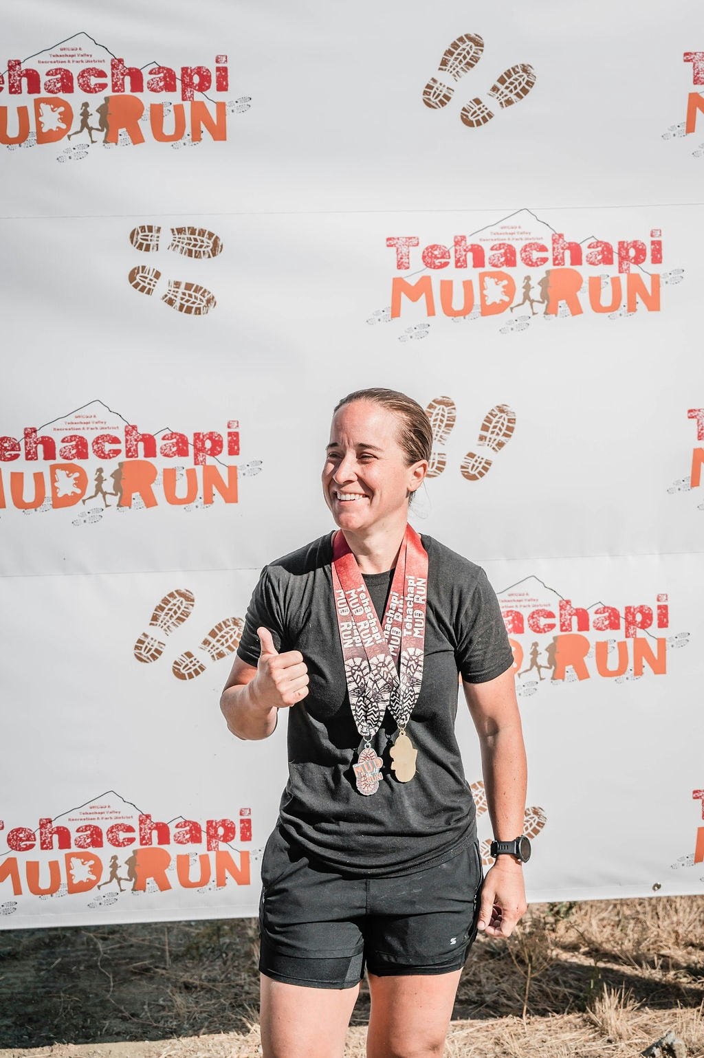 Person smiling, giving a thumbs up, wearing medals in front of Tehachapi Mud Run backdrop.