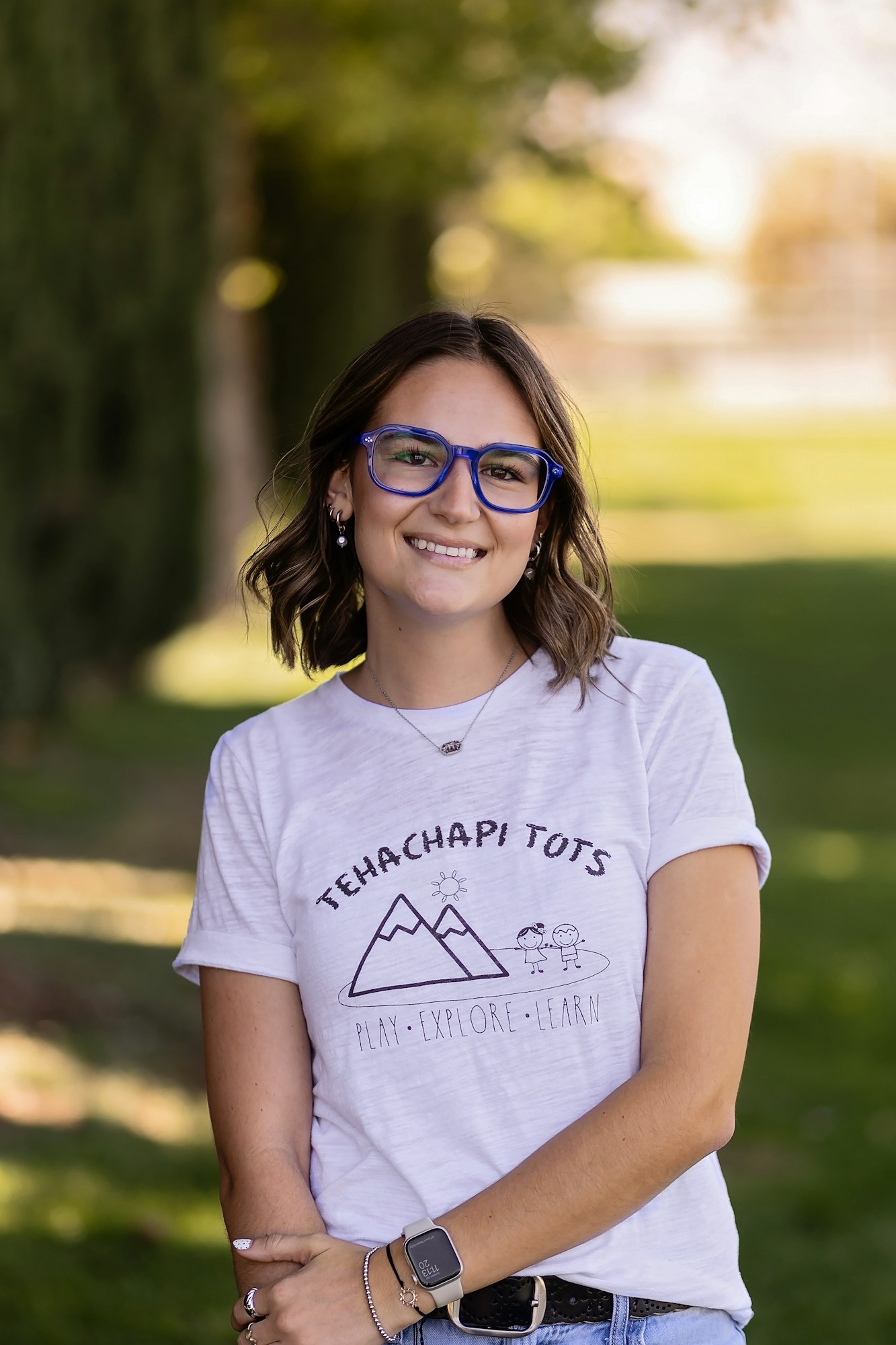 A person wearing a "Tehachapi Tots" T-shirt and blue glasses, standing outdoors.