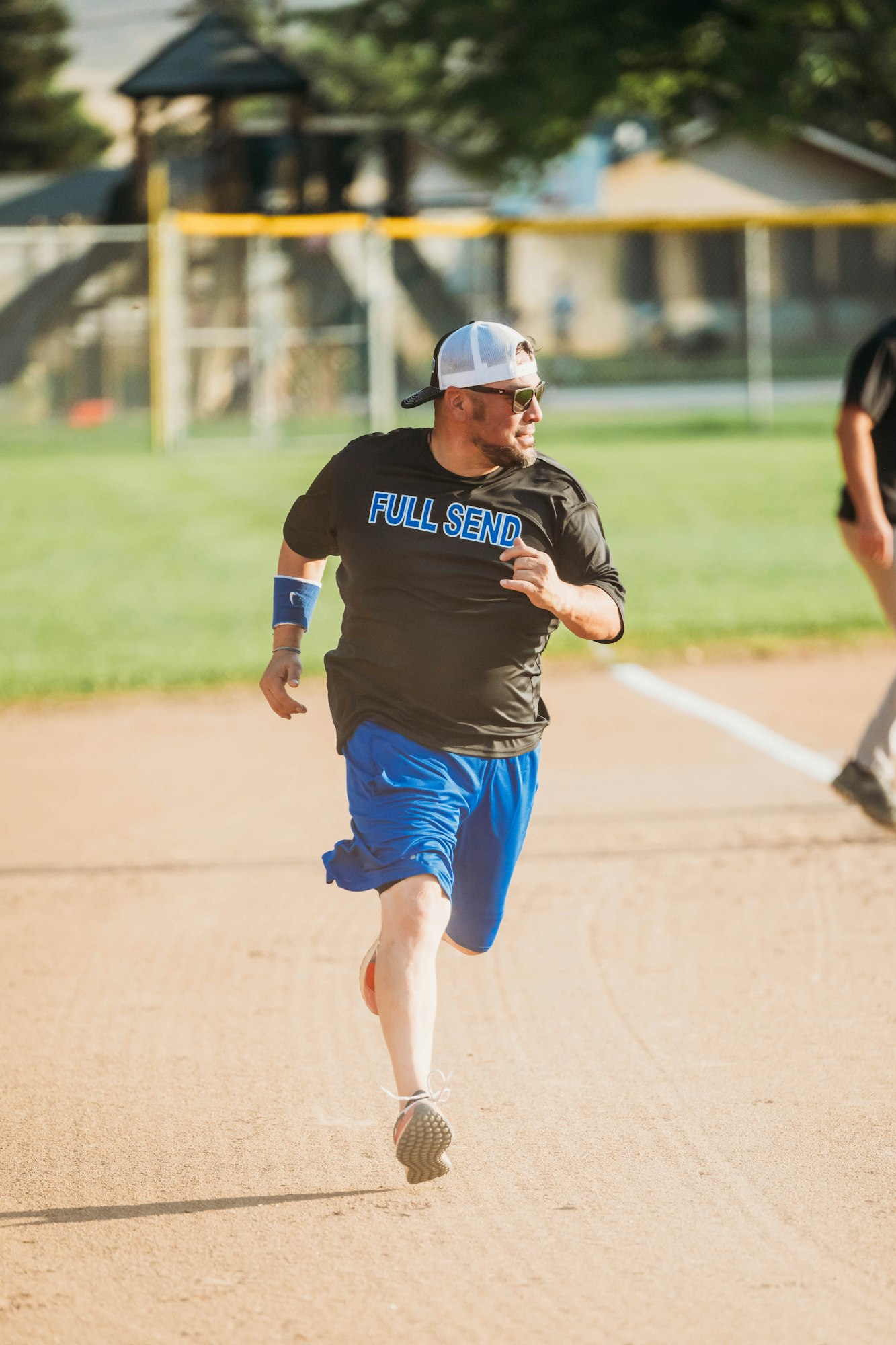 A person running on a softball field, wearing a "FULL SEND" t-shirt, sunglasses, a cap, and sports attire.