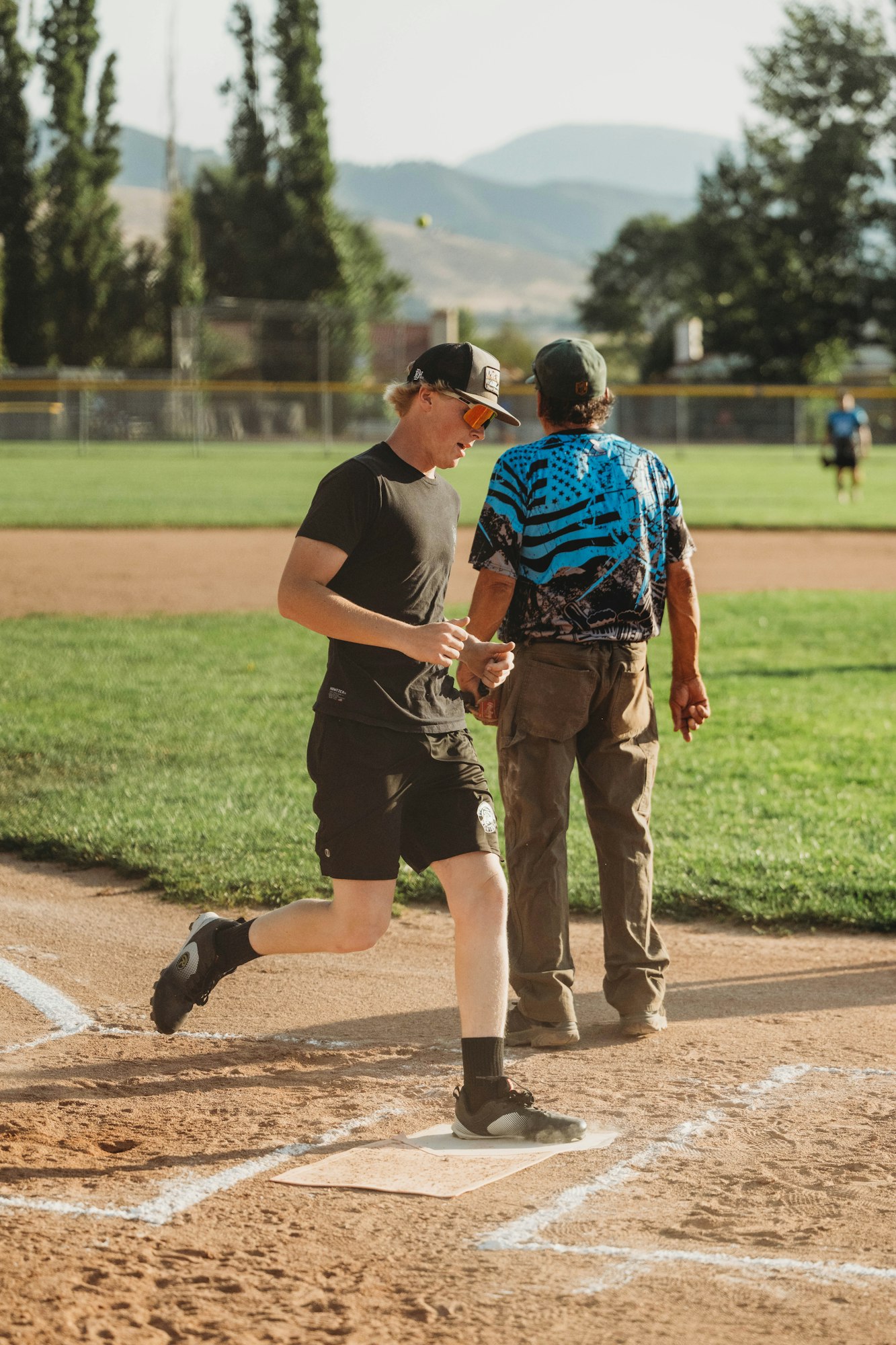 A player touches base in a baseball game with an umpire nearby; mountains and trees in the background.