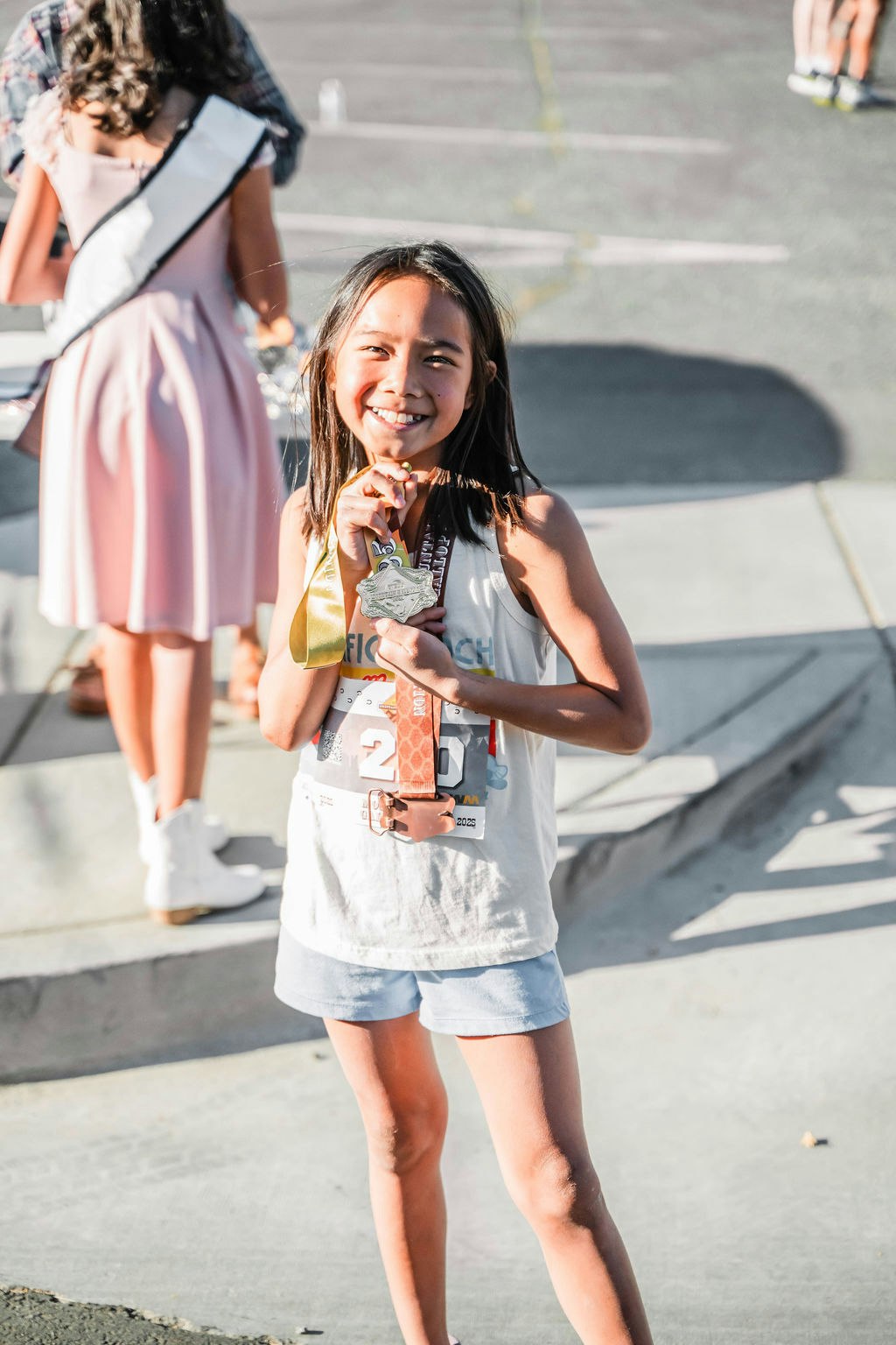 A smiling child holding a medal, wearing shorts and a tank top, standing outdoors.