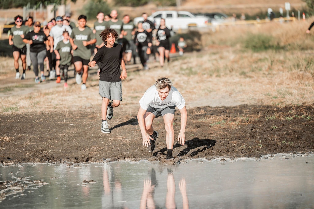 A group of people running; person in front is crouched near a muddy puddle, appearing to splash.