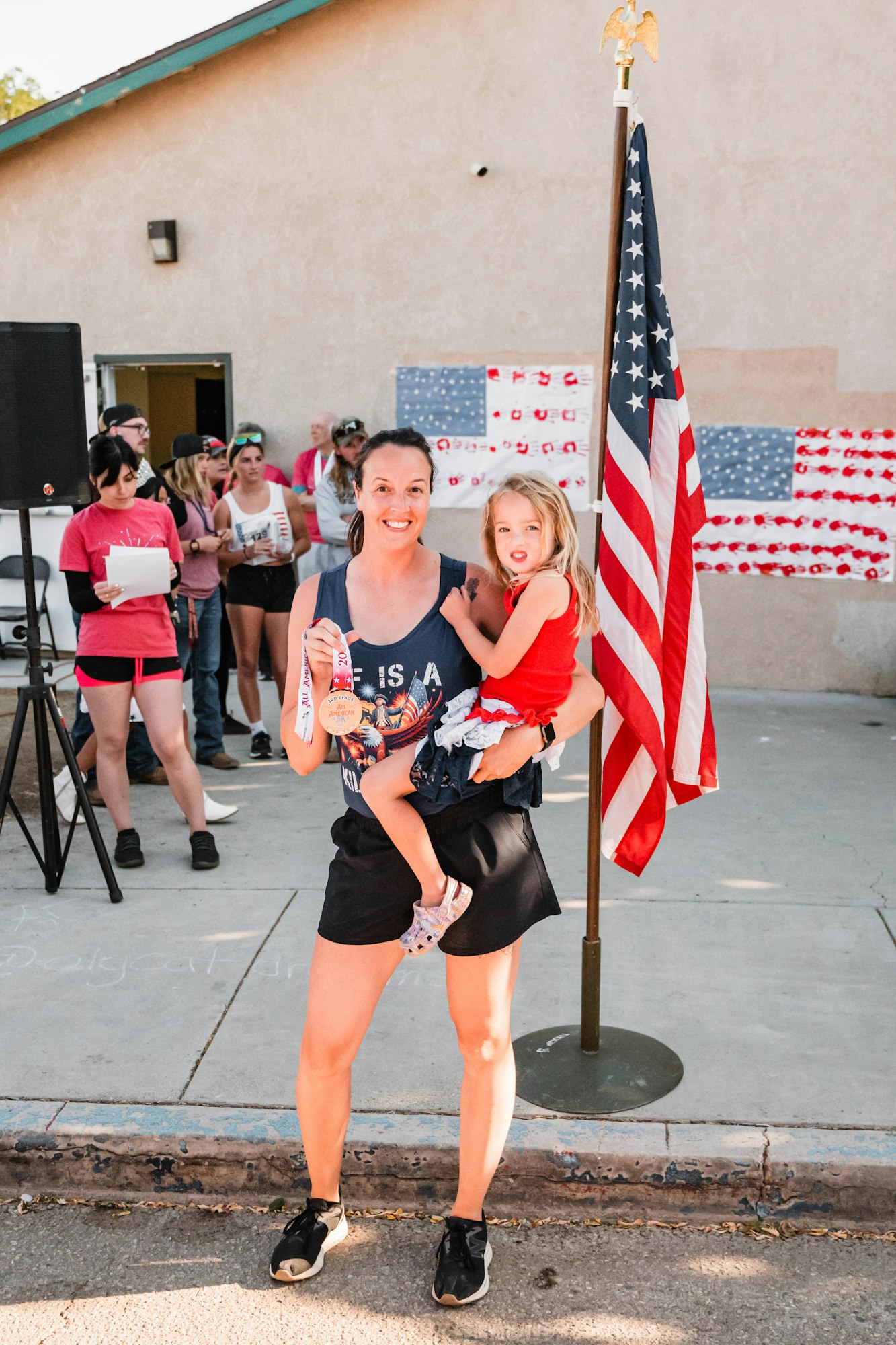 Smiling woman holding child near a U.S. flag, surrounded by people and wall decorations.