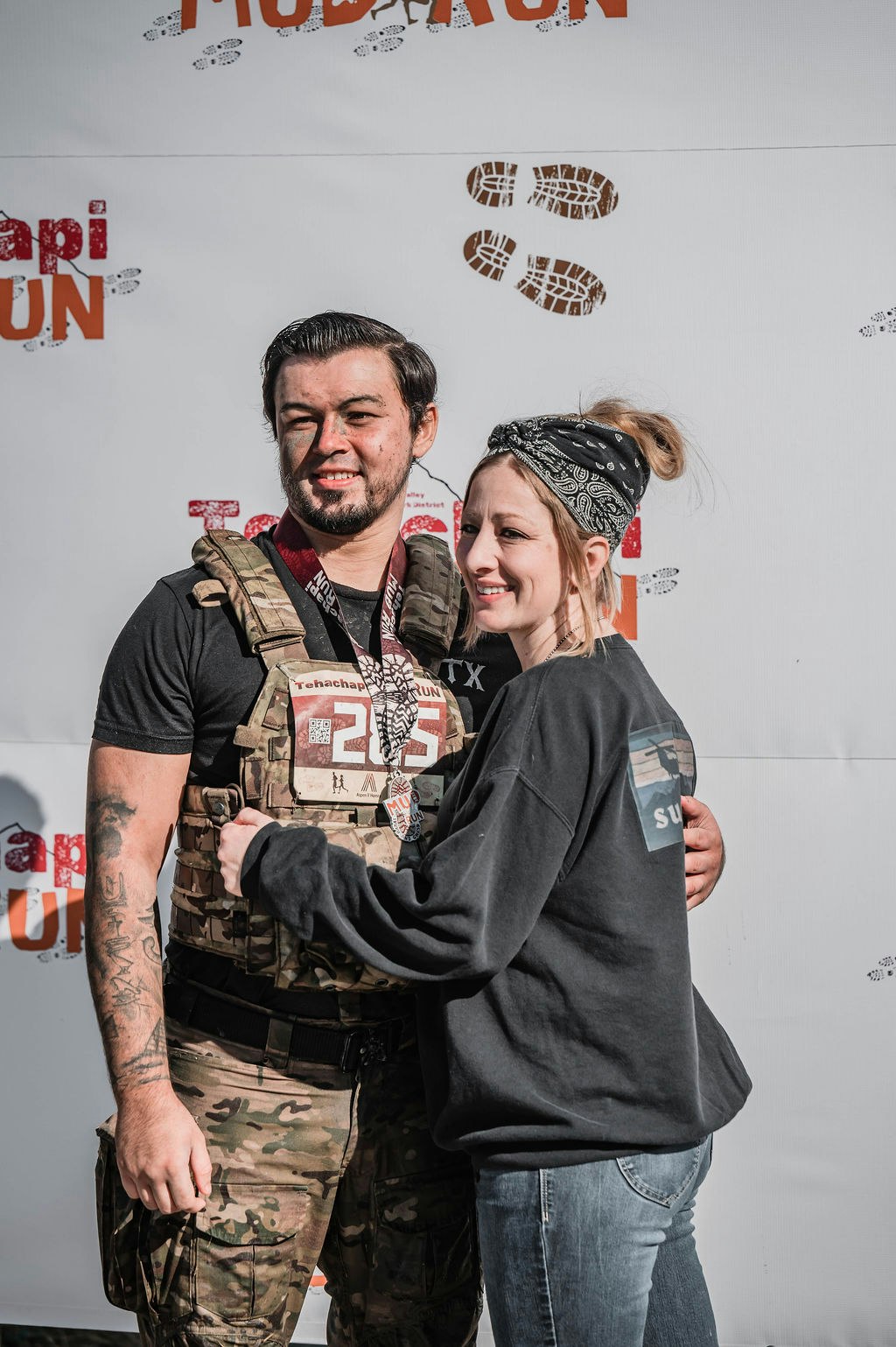 Two people posing together at a mud run event. One is wearing camouflage gear and a bib number.