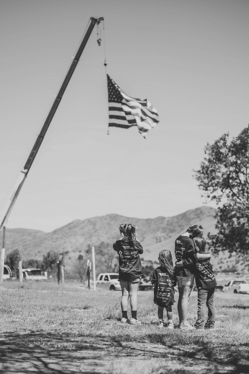 A group of people, likely a family, is looking up at a large US flag hanging from a crane in an open field, in black and white.