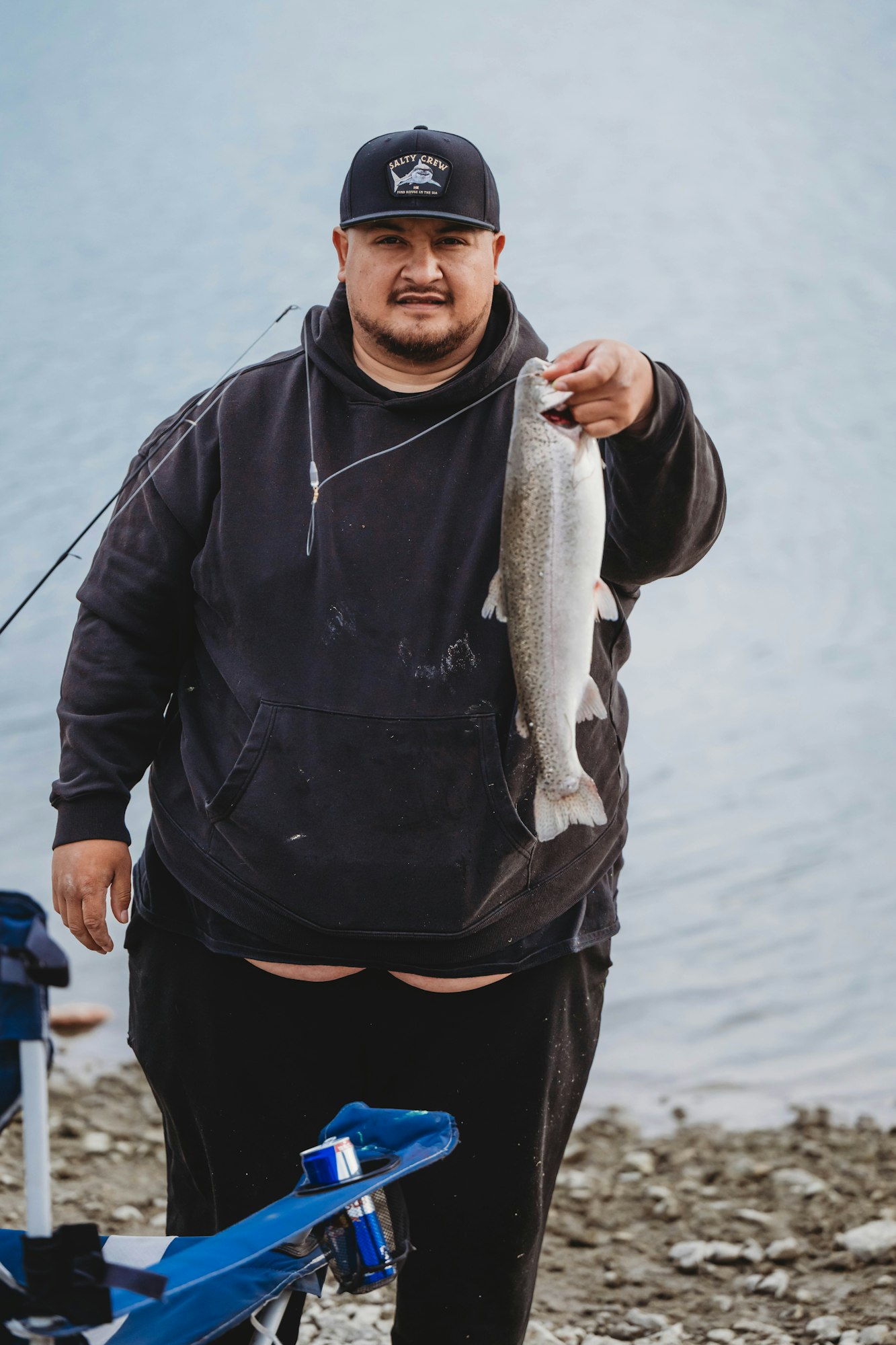 A person in black holds a fish by the water, wearing a cap and hoodie, standing next to a chair with items on it.
