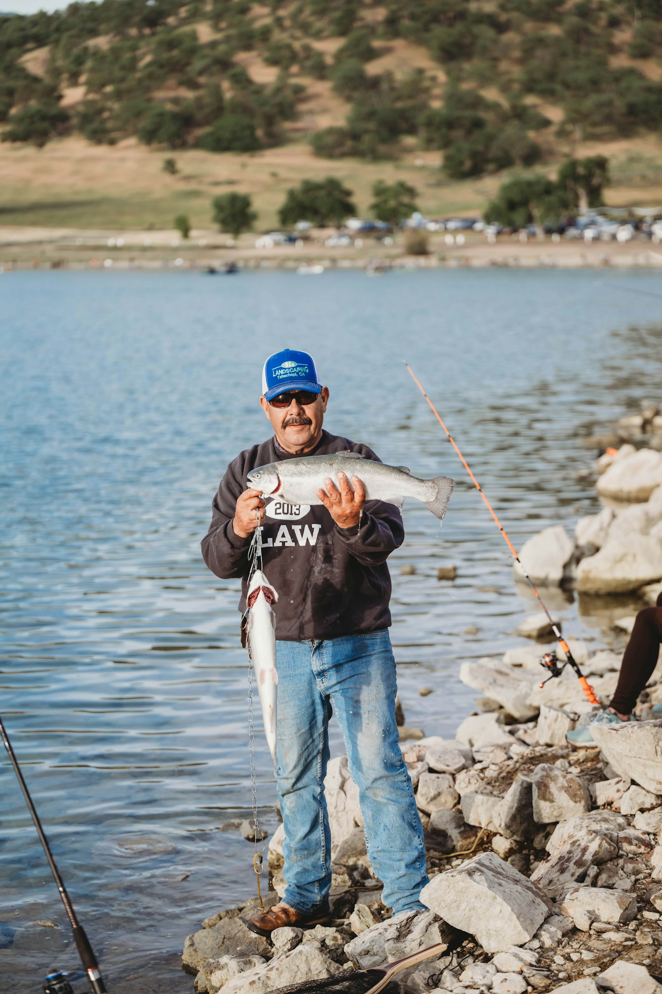 A person holds two fish by a lake, standing on rocks, wearing a blue cap and sunglasses. Fishing rods are visible nearby.