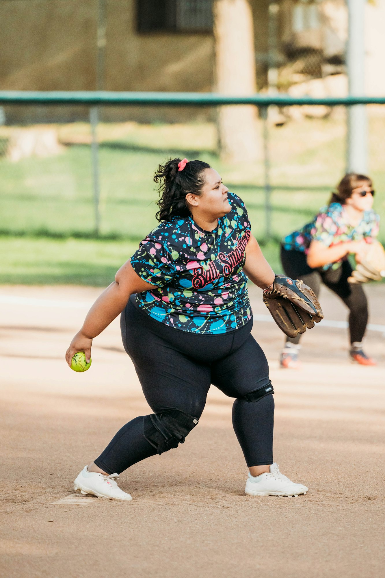A softball player in a colorful jersey prepares to pitch on a sunny day. Another player is seen in the background.