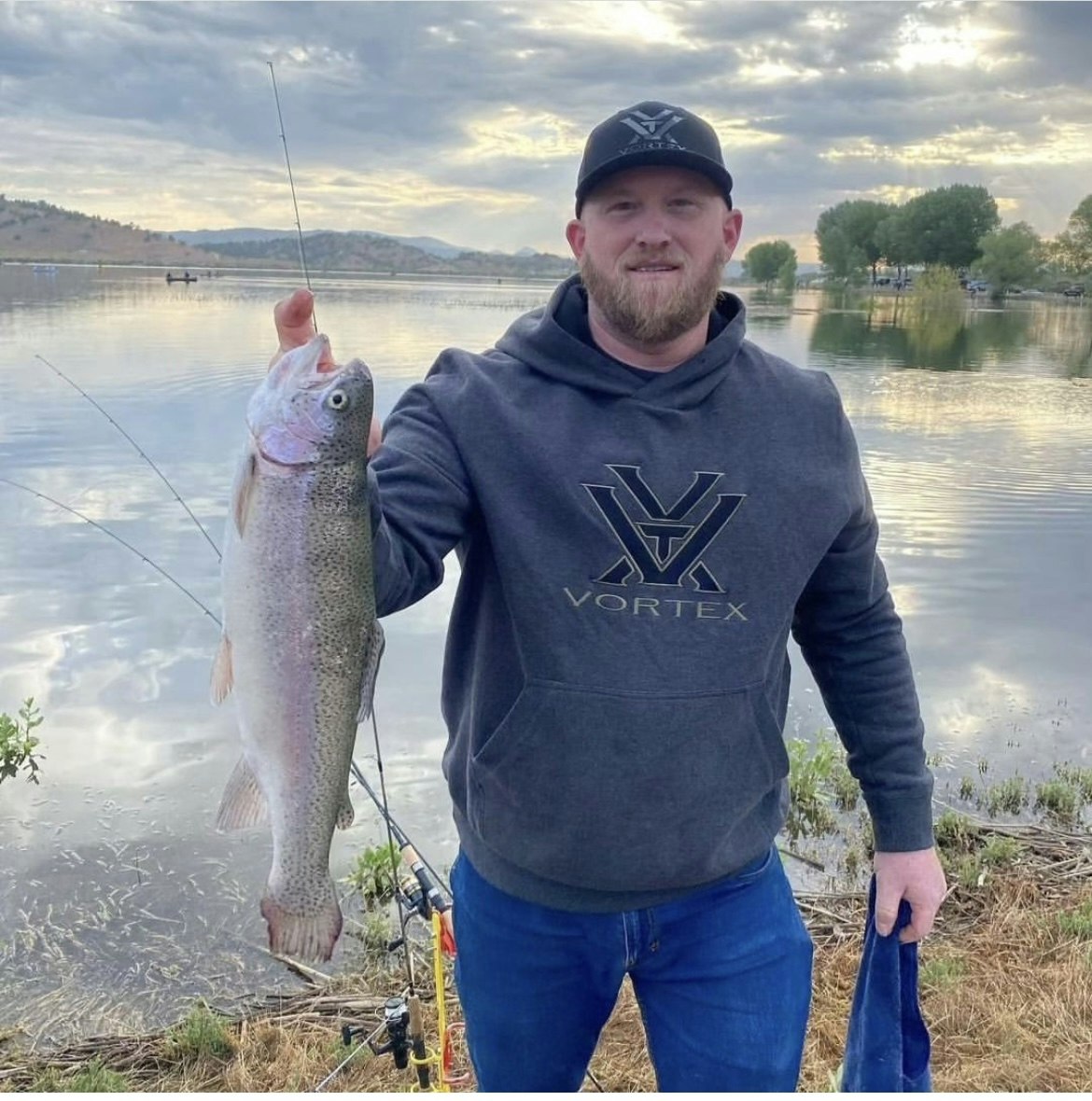 A man proudly holds a large fish by a lake with fishing rods nearby.
