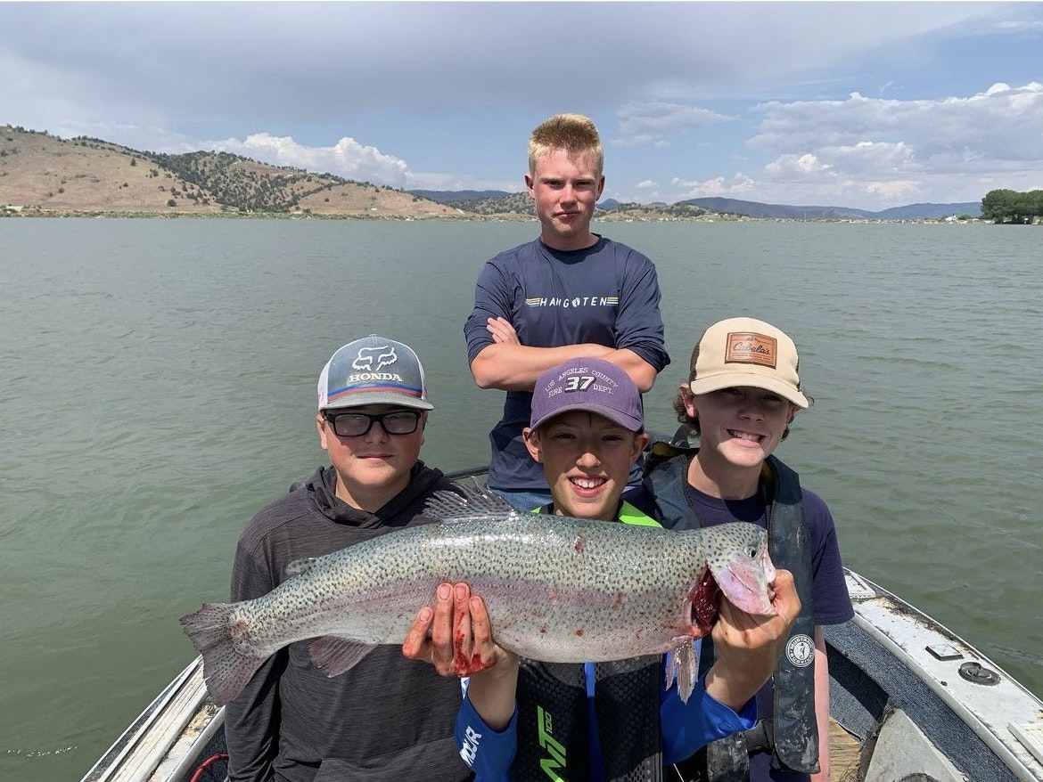 Three youths holding a large fish in a boat on a lake with hills in the background.