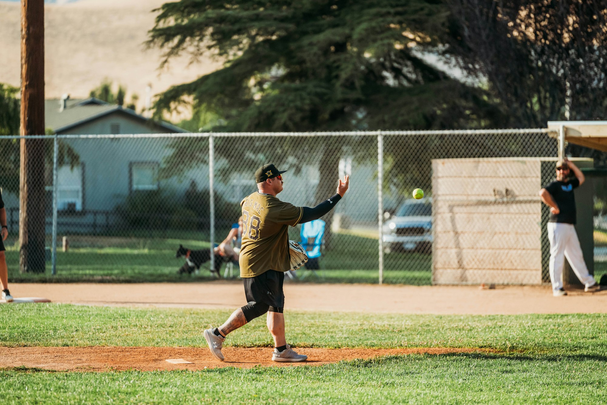A person pitches a softball on a sunny day at a park, with another player in the background.