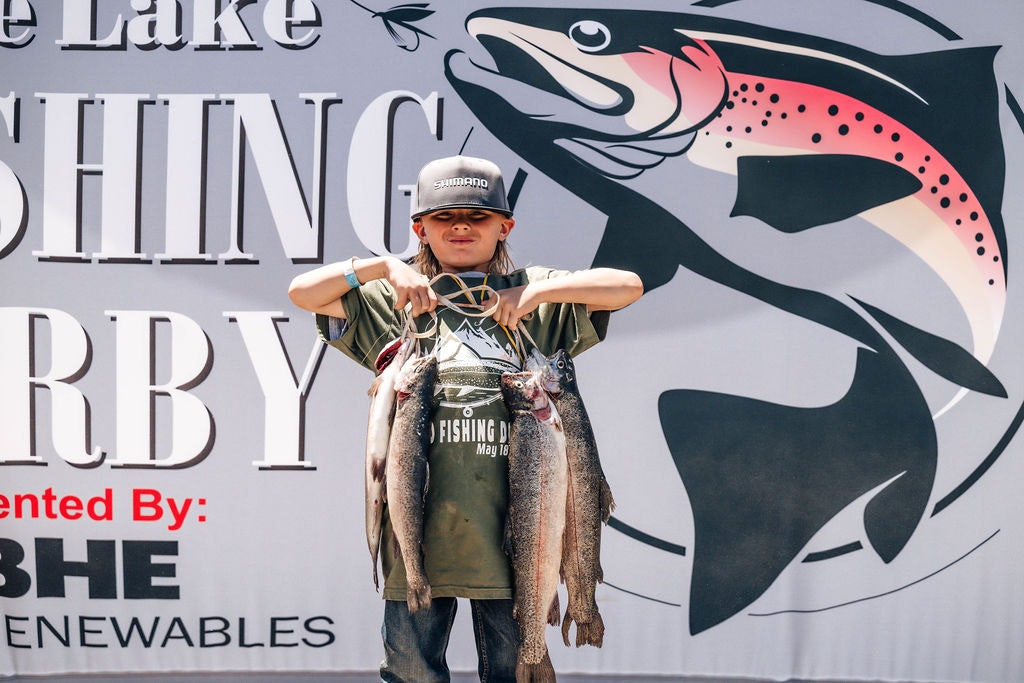 A kid holding two large fish in front of a banner for a fishing derby.