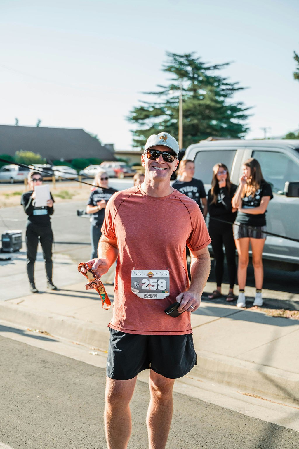 A man in a red shirt with a race bib, holding a medal, standing outdoors with a group of people behind him.