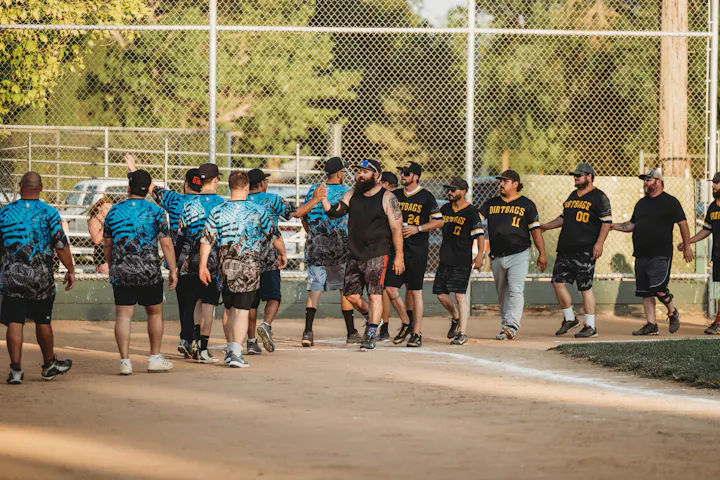 Two softball teams shaking hands after a game on a dirt field, with a chain-link fence and trees in the background.