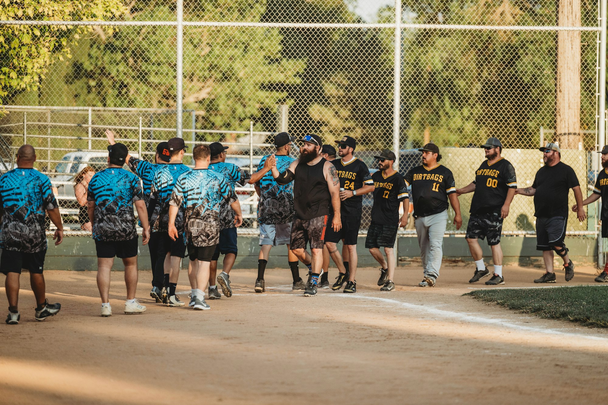 Two softball teams shaking hands after a game on a dirt field, with a chain-link fence and trees in the background.
