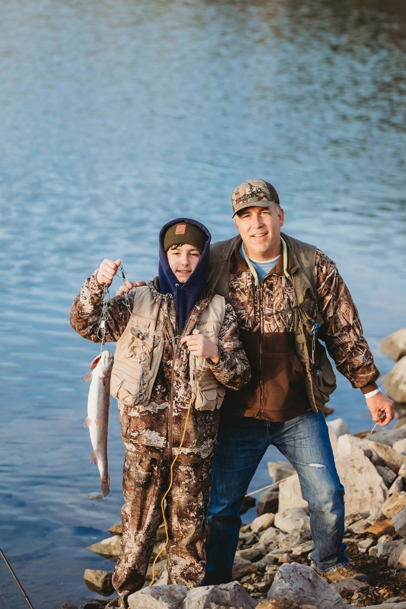 Two people in camouflage clothing by a lake; one holds a fish.