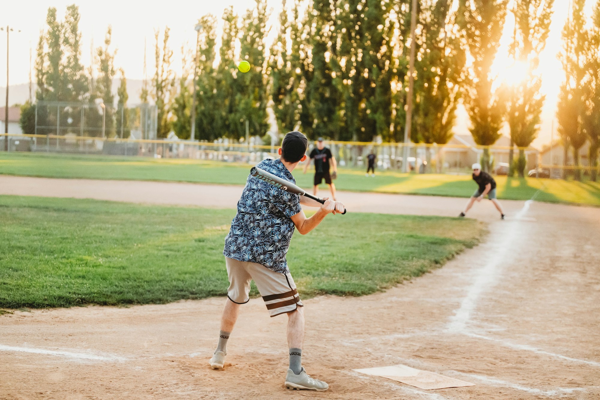 A person in a floral shirt prepares to bat during a baseball game at sunset, with other players in the background.