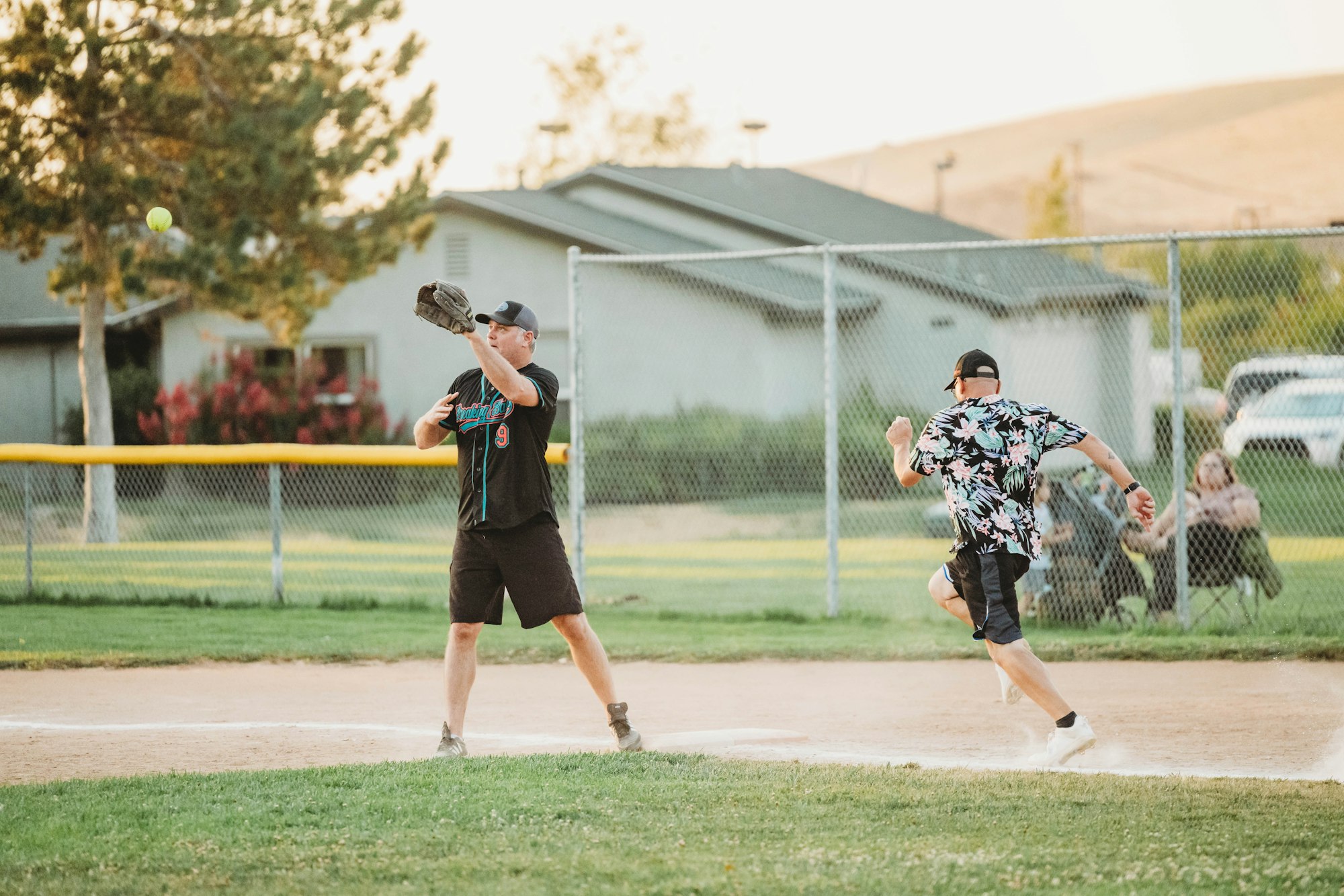 A baseball player prepares to catch a ball while another runs towards home plate in a sunny park setting.